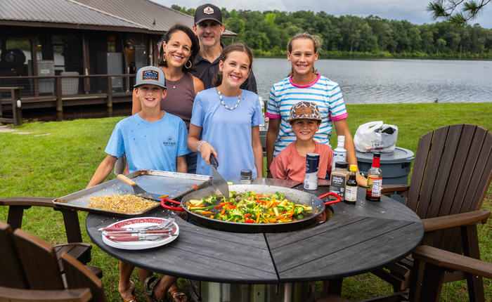 Family gathered outdoors by a lake around a round charcoal grill table with grilled vegetables.