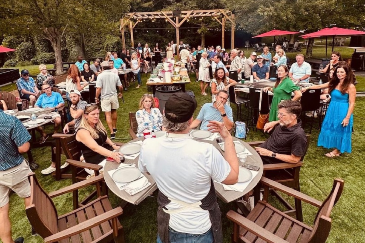 Large outdoor gathering with people seated around round dining tables on grass, set for grilling.