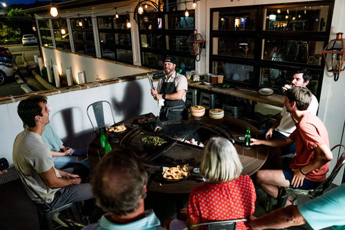 Group gathered at night around a large outdoor grill table enjoying food and drinks