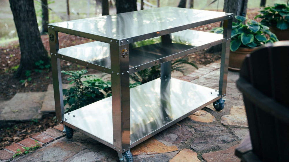Stainless steel outdoor prep table with wheels on a stone patio, surrounded by plants
