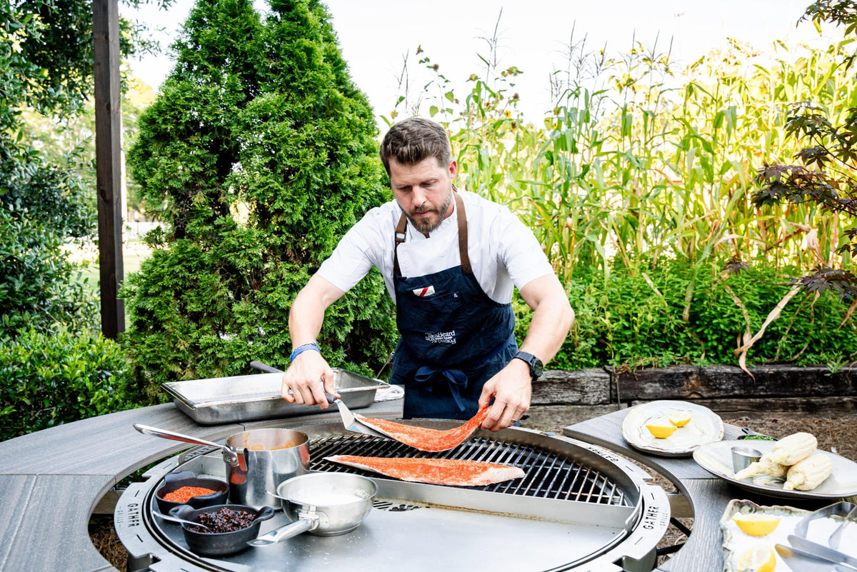 Chef grilling seasoned fish fillets on a round outdoor charcoal grill with cooking ingredients nearby
