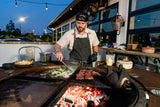 Man grilling steak and vegetables on outdoor rotating charcoal grill at night